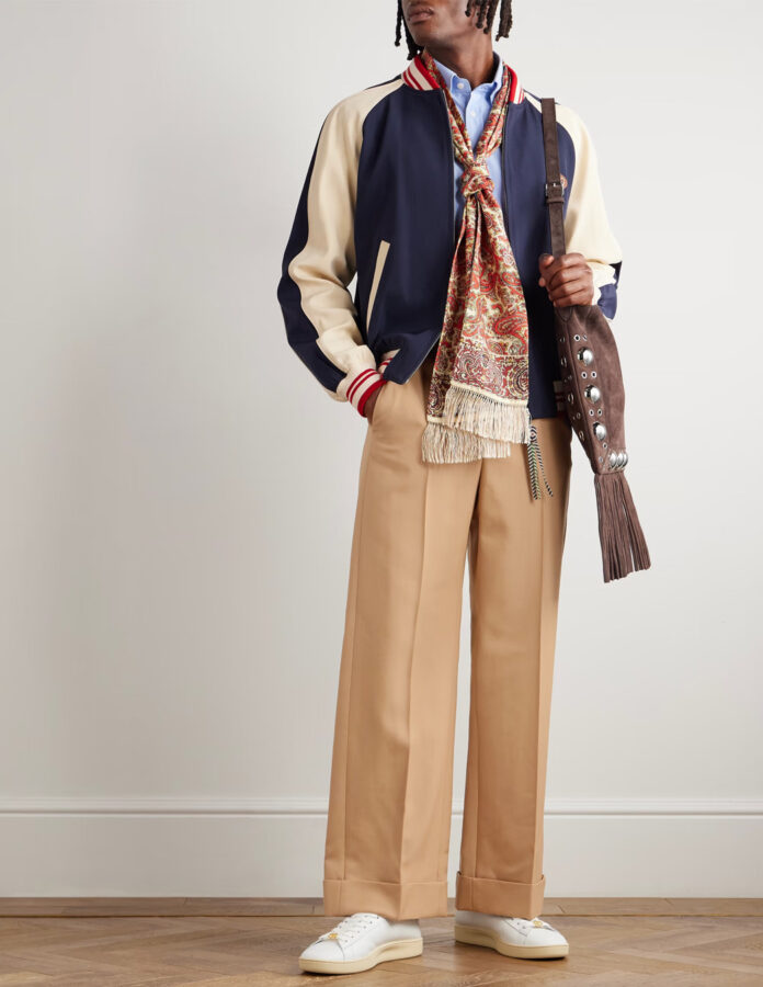 hombre vestido con pantalón beige conjuntado con camisa, chaqueta varsity, zapatillas blancas clásicas y accesorios.