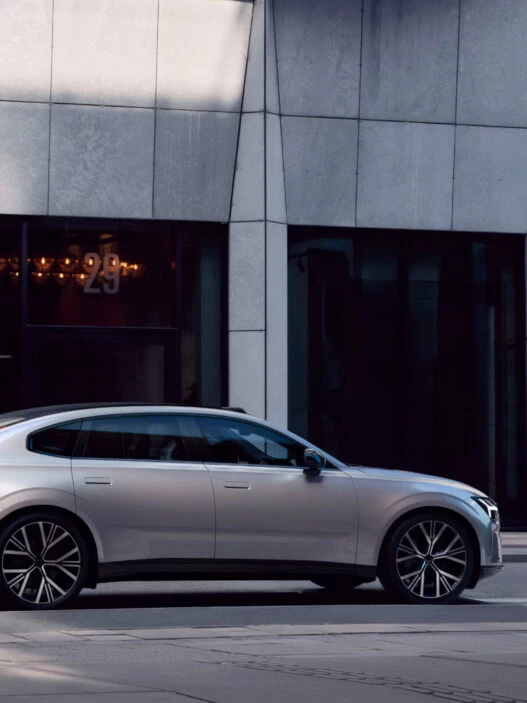 Volvo ES90 2025 en color plateado circulando por una calle urbana con edificios modernos