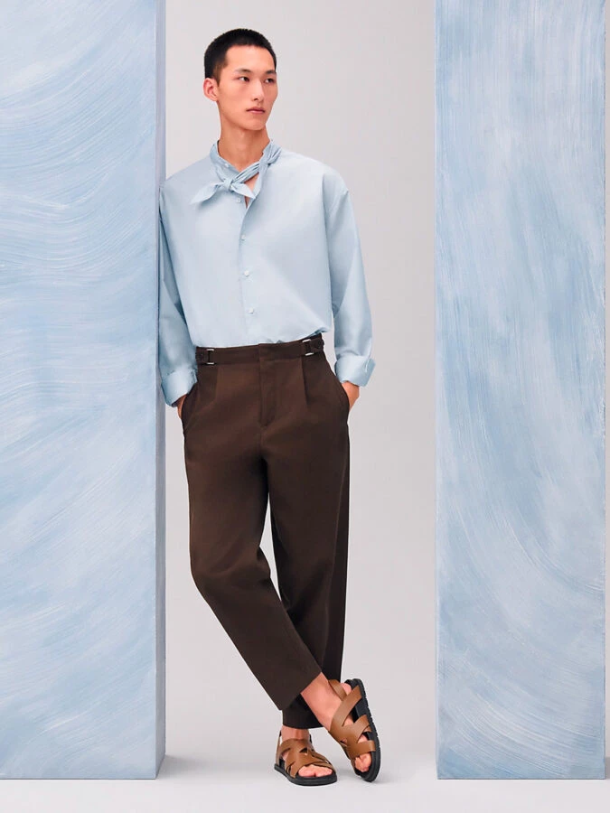 Look de Hermès con camisa celeste, pañuelo al cuello, pantalón marrón y sandalias de cuero