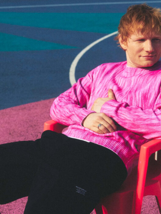 Ed Sheeran sentado en una silla roja con jersey rosa en una pista multicolor