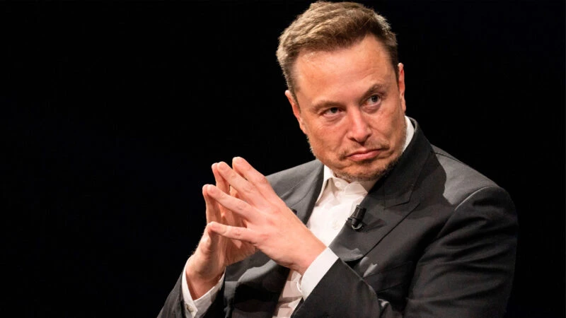 Elon Musk en un evento, con traje oscuro y manos entrelazadas