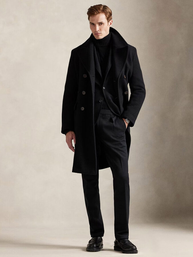 Cómo combinar el pantalón negro: modelo masculino vestido con jersey de cuello alto negro y abrigo largo negro con mocasines