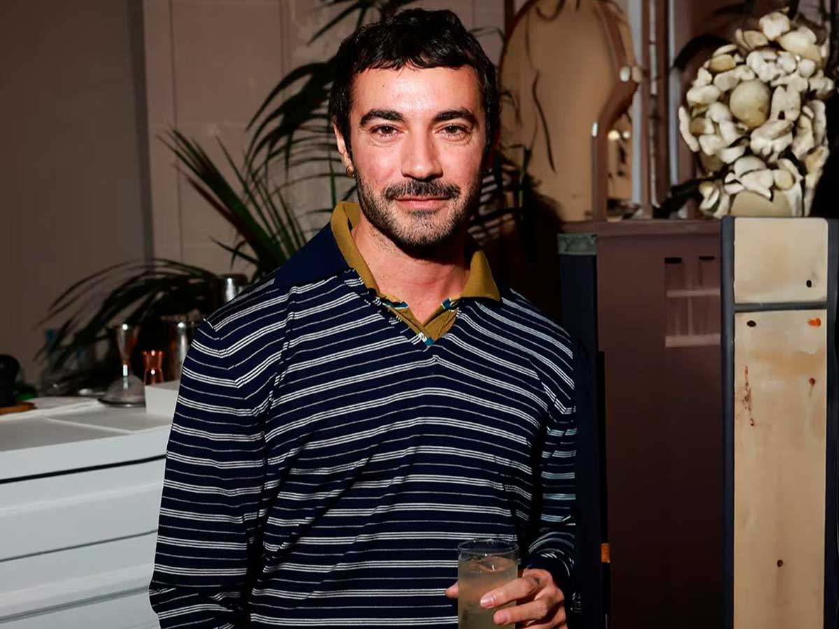 Retrato del diseñador Dario Vitale en el que viste un polo de rayas blancas finas sobre un fondo azul. El diseñador agarra un cóctel en la mano