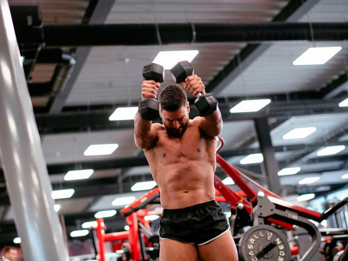 hombre sin camiseta entrenando hombro con mancuernas en un gimnasio