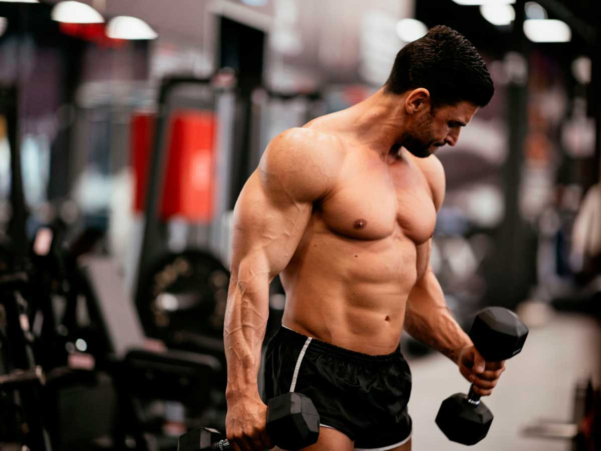 hombre en un gimnasio entrenando pull de biceps con mancuernas en un gimnasio y sin camiseta