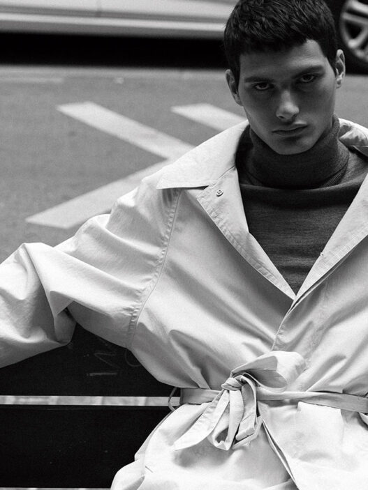 El modelo Alejo Humanes vestido con un jersey de cuello alto y una gabardina y sentado en un banco