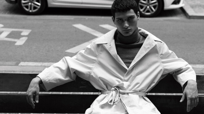 El modelo Alejo Humanes vestido con un jersey de cuello alto y una gabardina y sentado en un banco
