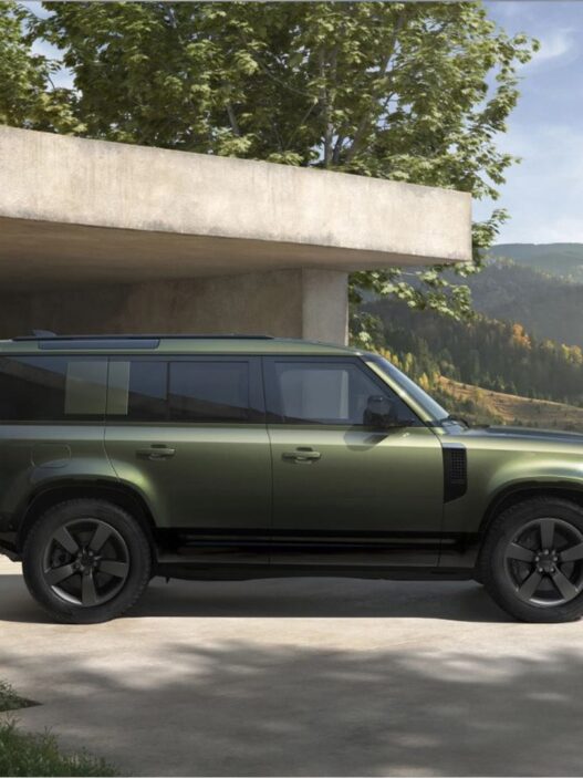 Coche Land Rover Defender 110 verde aparcado en la puerta de una casa de campo muy moderna con vistas a un campo verde iluminado por la luz de un día soleado.