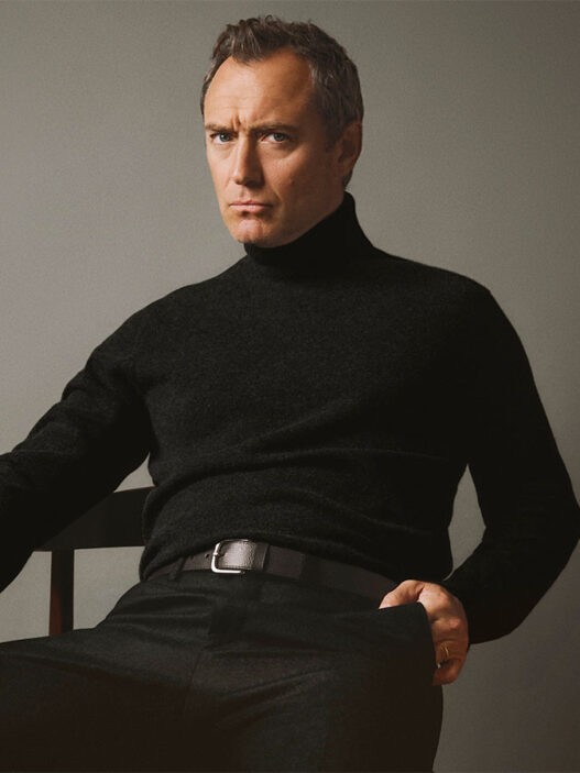 Jude Law en una imagen de la nueva campaña de Emidio Tucci otoño 2025 en la que aparece sentado en una silla con un jersey de cuello alto negro y semblante serio