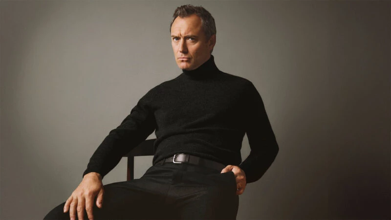Jude Law en una imagen de la nueva campaña de Emidio Tucci otoño 2025 en la que aparece sentado en una silla con un jersey de cuello alto negro y semblante serio