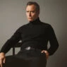 Jude Law en una imagen de la nueva campaña de Emidio Tucci otoño 2025 en la que aparece sentado en una silla con un jersey de cuello alto negro y semblante serio
