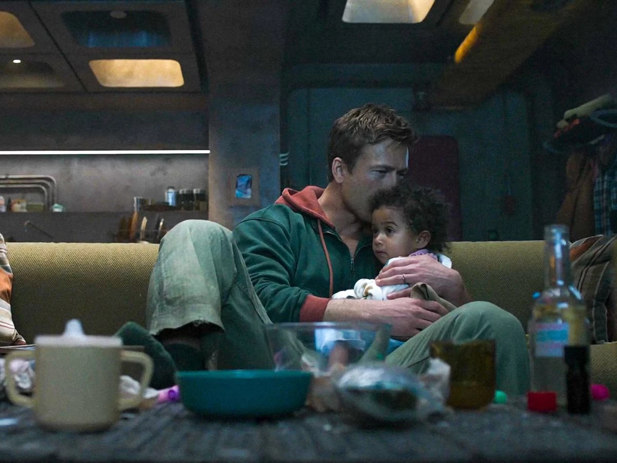 Escena de la película The Running Man Stephen King en la que se ve a su protagonista dándole un beso en la cabeza a una niña pequeña, ambos están sentados en un sofá