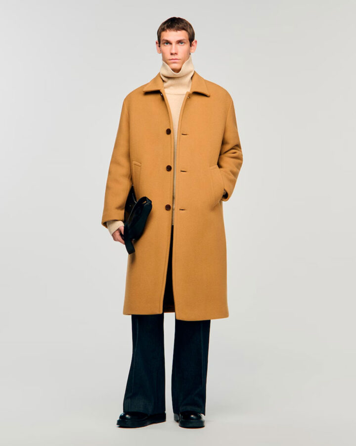 Modelo masculino de unos veinti-pocos años vestido con abrigo largo por debajo de la rodilla, color camel claro, un pantalón ancho y un jersey de cuello alto color claro.