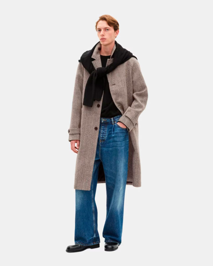 modelo joven vestido con un abrigo largo por debajo de la rodilla de espiga y color marrón, combinado con pantalones vaqueros y botas negras. El modelo lleva ta,bién un jersey negro al hombro