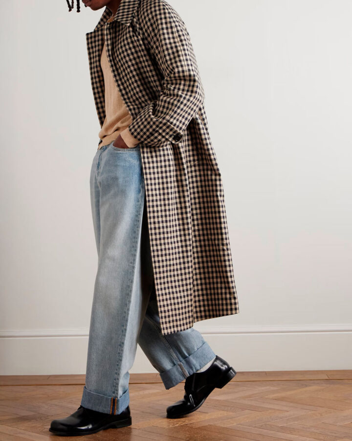 modelo jovencito, de unos veinte años y piel oscura, vestido muy cool con un abrigo largo de pata de gallo color beige, marrón y negro, con unos pantalones vaqueros de pernera ancha y cintura alta, un jersey de punto color piel, y unos zapatos de cordones negros.