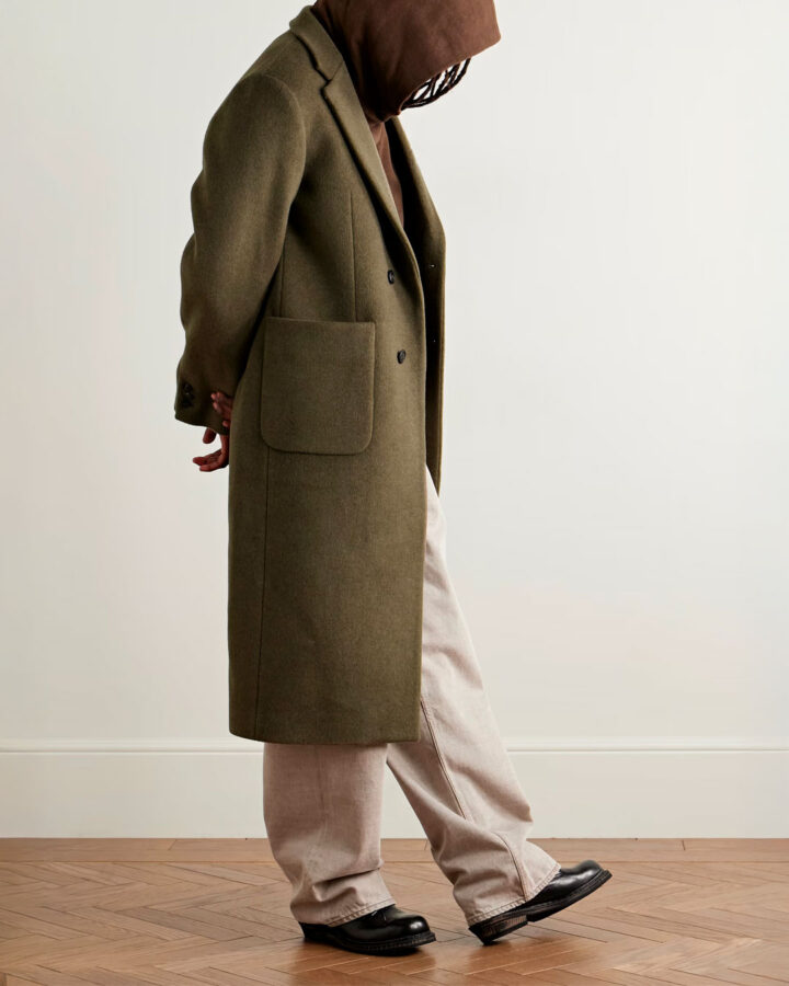 Modelo vestido con un abrigo largo verde por debajo de la rodilla, un pantalón denim ancho y una sudadera marrón chocolate