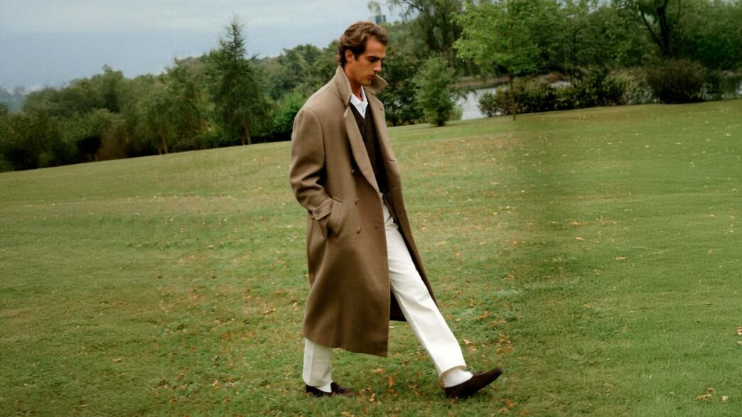 chico de unos veinte años vestido muy cool con un abrigo largo de lana color marrón combinado con unos pantalones blancos y un jersey de pico. El chico camina por mitad de un campo verde en una mañana fría mañana de otoño