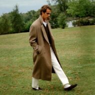 chico de unos veinte años vestido muy cool con un abrigo largo de lana color marrón combinado con unos pantalones blancos y un jersey de pico. El chico camina por mitad de un campo verde en una mañana fría mañana de otoño