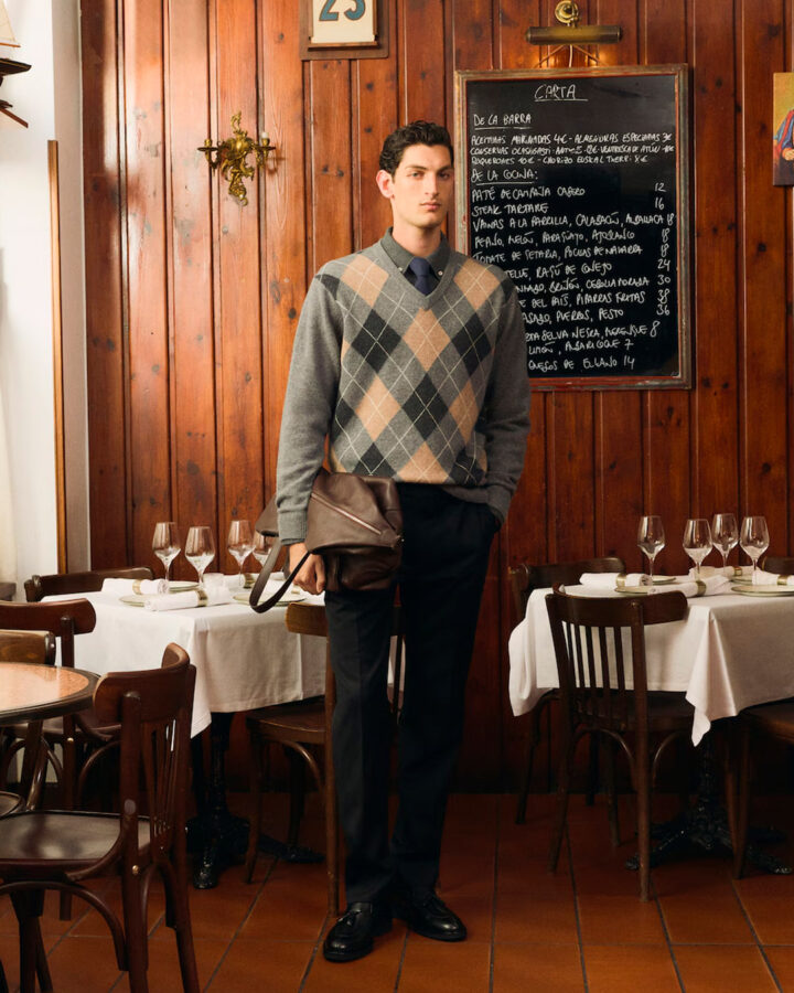 Hombre con jersey de rombos gris de Emidio Tucci combinado con camisa azul y pantalones de pinzas en un restaurante clásico de Madrid.
