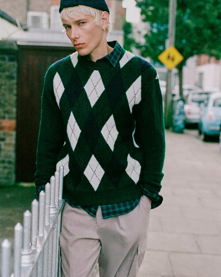 Hombre con jersey verde de rombos blancos y burdeos de Sfera, camisa de cuadros y pantalones beige en un look urbano.