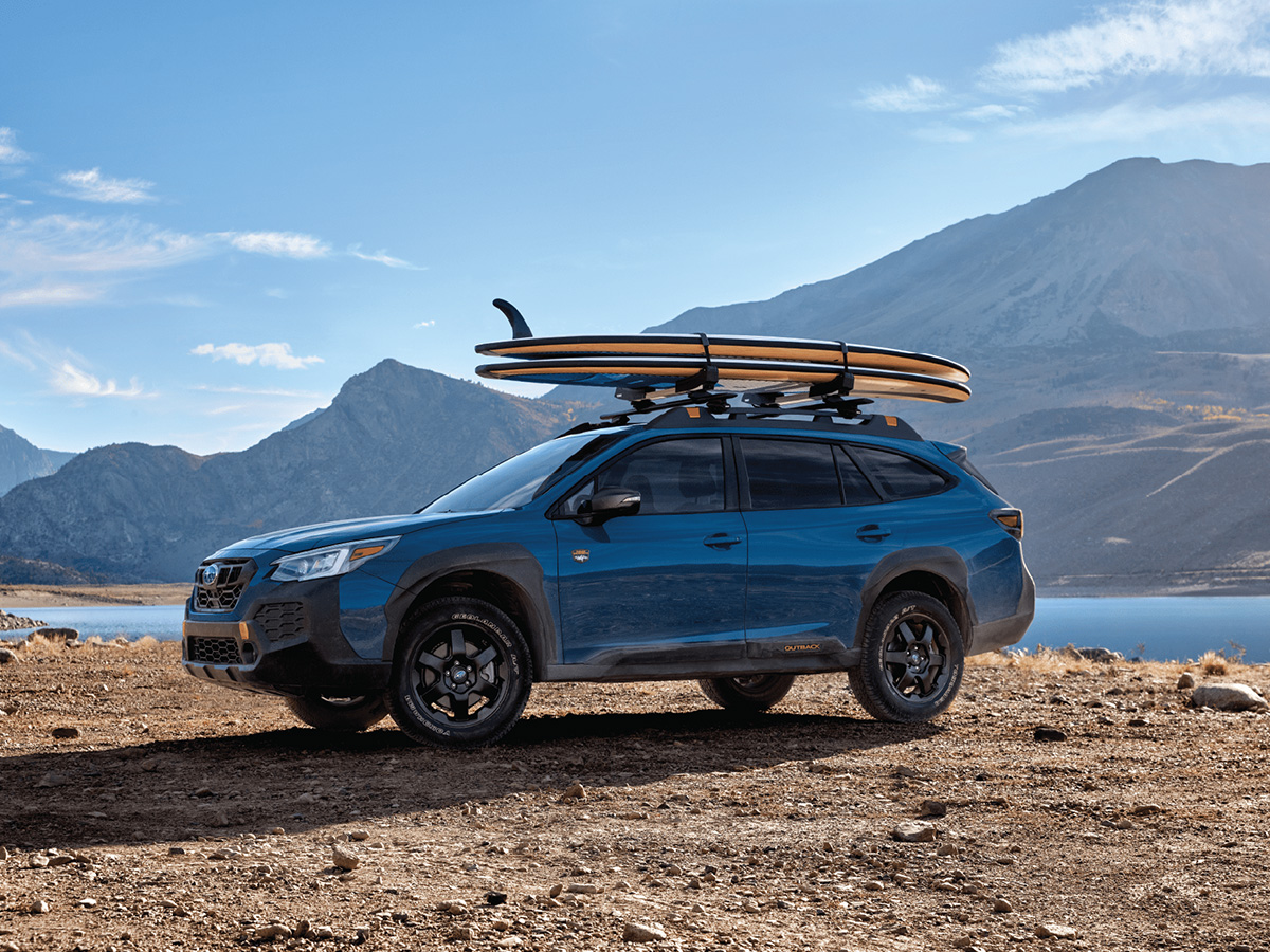 Coche Subaru Outback en color azul con unas tablas de surf enganchadas en el techo. El coche está frente a una playa.
