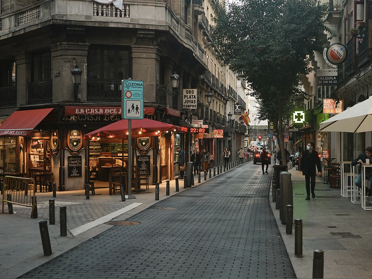 Fotografía de una de las calles del centro de Madrid, posiblemente barrio de Malasaña, con gente andando por la acera y escaparates encendidos