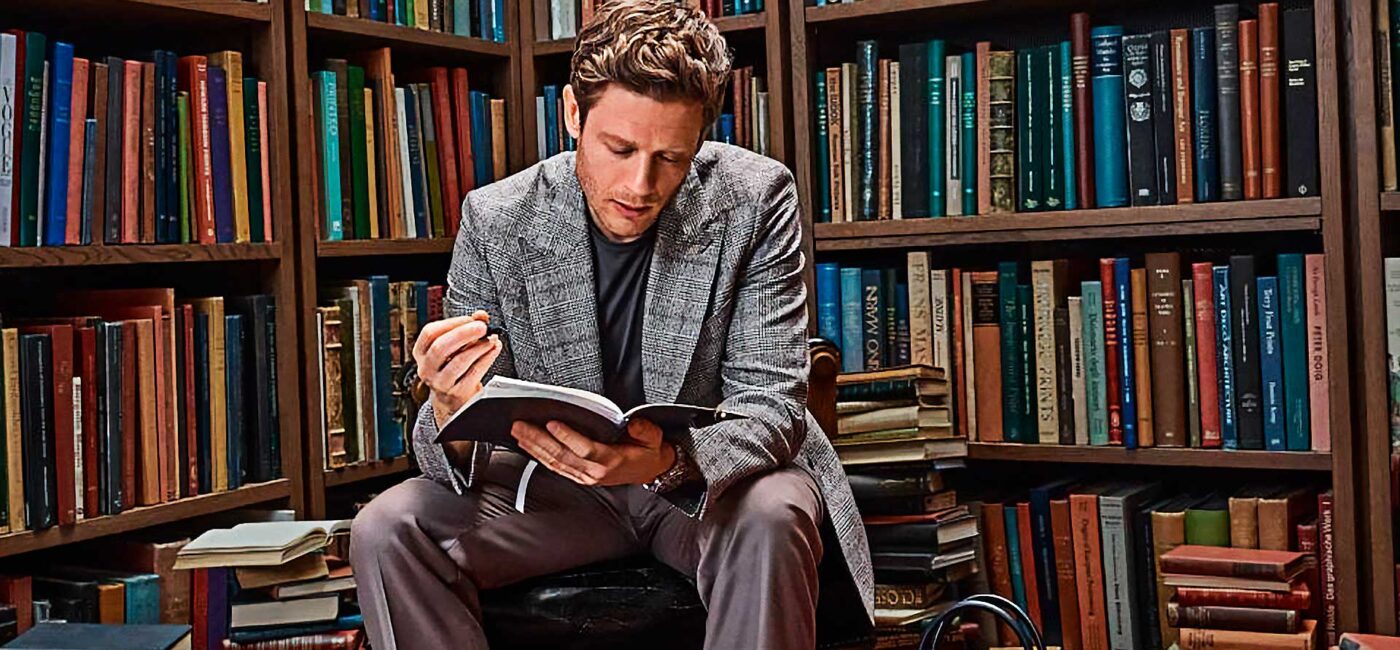 Hombre de unos 35 años, vestido muy elegante con un abrigo de espiga gris, y un jersey, leyendo un libro en una biblioteca de estilo antiguo repleta de libros. El hombre está sentado muy concentrado leyendo un libro. Al rededor se ven libros apilados y ordenados en estanterías.