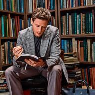Hombre de unos 35 años, vestido muy elegante con un abrigo de espiga gris, y un jersey, leyendo un libro en una biblioteca de estilo antiguo repleta de libros. El hombre está sentado muy concentrado leyendo un libro. Al rededor se ven libros apilados y ordenados en estanterías.
