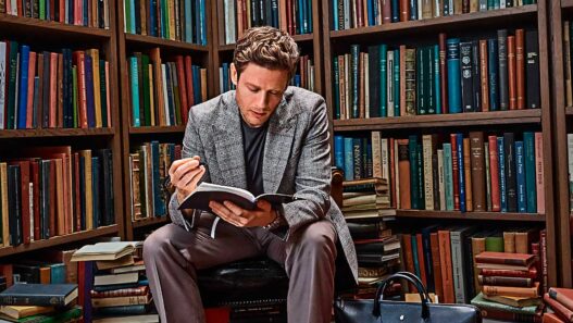 Hombre de unos 35 años, vestido muy elegante con un abrigo de espiga gris, y un jersey, leyendo un libro en una biblioteca de estilo antiguo repleta de libros. El hombre está sentado muy concentrado leyendo un libro. Al rededor se ven libros apilados y ordenados en estanterías.
