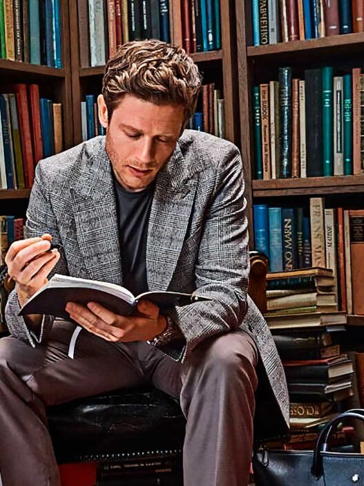 Hombre de unos 35 años, vestido muy elegante con un abrigo de espiga gris, y un jersey, leyendo un libro en una biblioteca de estilo antiguo repleta de libros. El hombre está sentado muy concentrado leyendo un libro. Al rededor se ven libros apilados y ordenados en estanterías.
