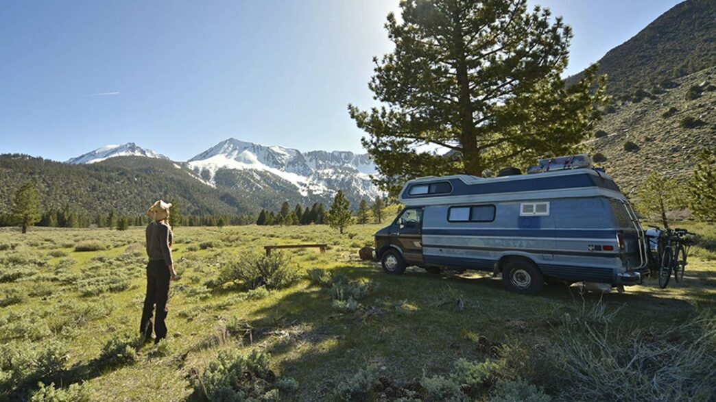 hombre en un bonito campo verde frente a su caravana de fondo se ven unas hermosas montañas nevadas en un día muy soleado