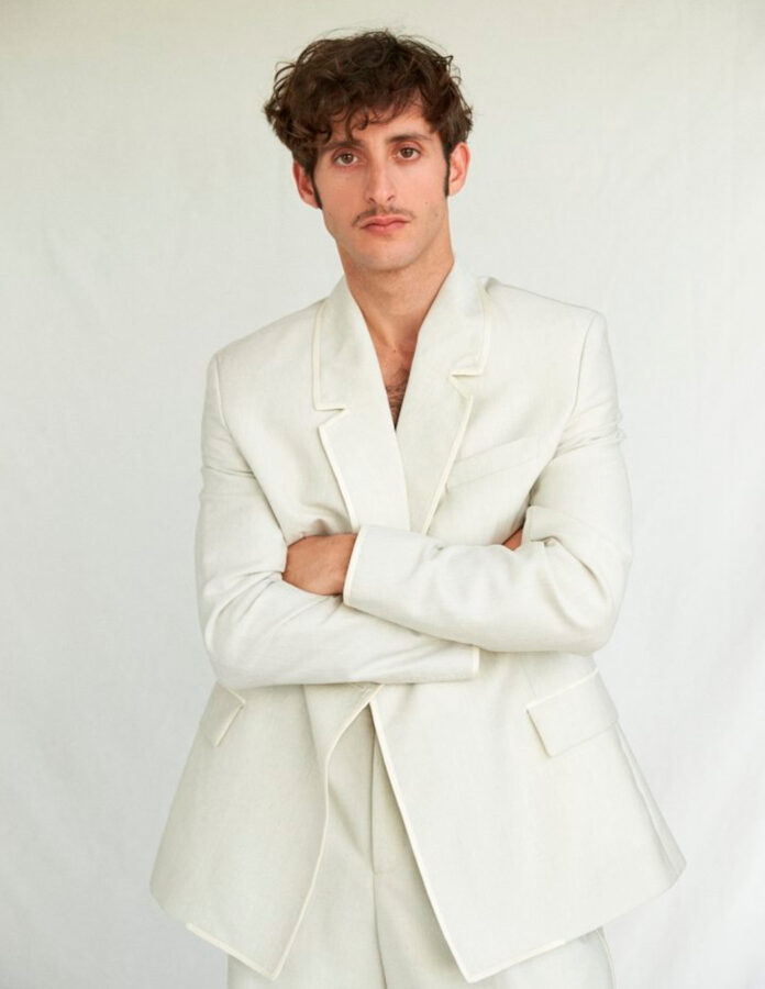 El actor Daniel Ibañez posa en un estudio, vestido con traje blanco y los brazos cruzados, rostro serio.