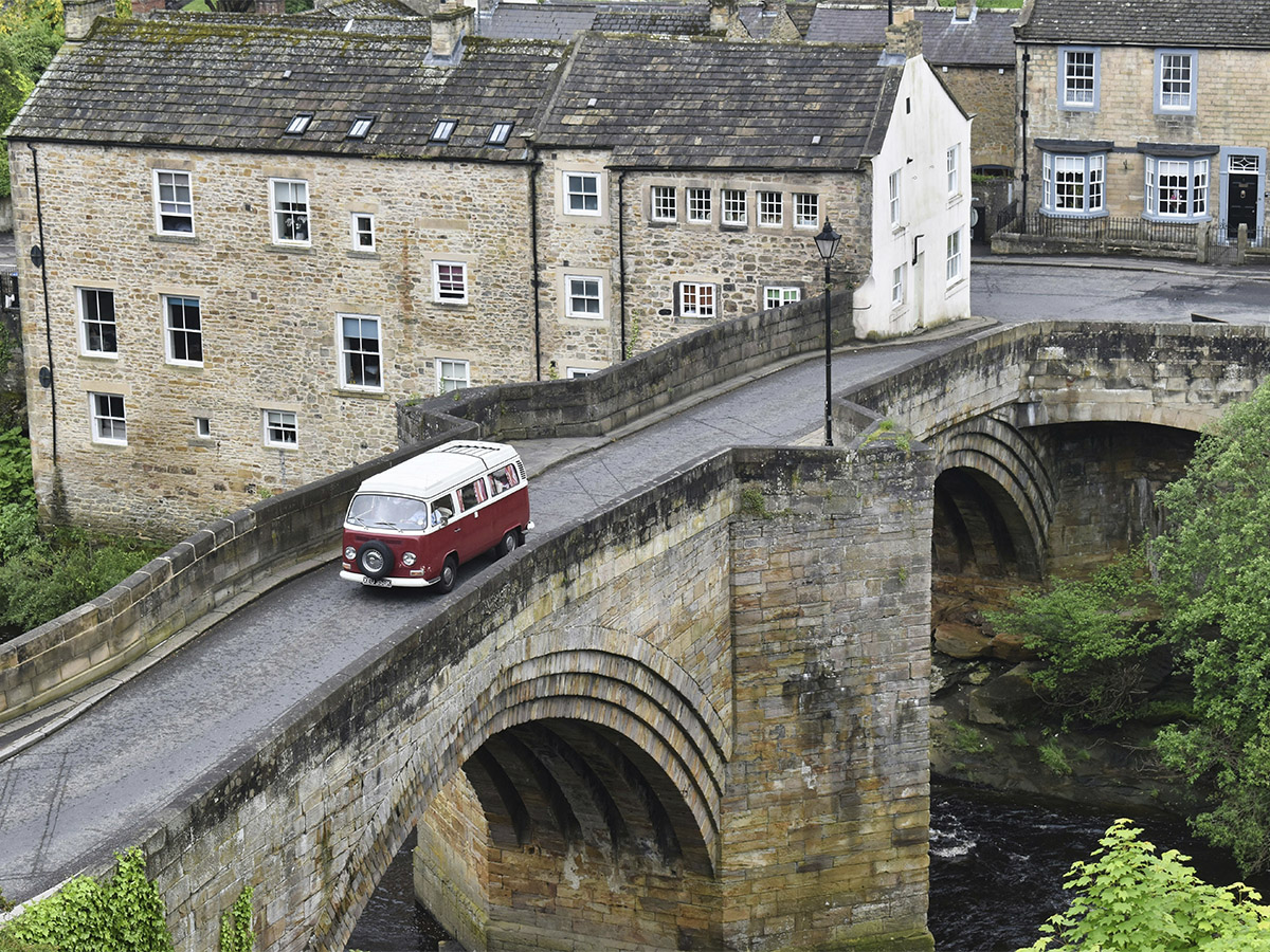 Vista aerea de una autocaravana roja cruzando un puente de piedra que llega a un pueblecito antiguo
