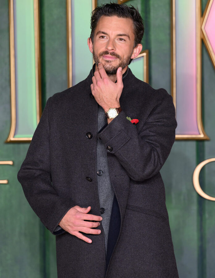 Jonathan Bailey con abrigo oscuro y reloj OMEGA Seamaster en la alfombra amarilla de Wicked: For Good