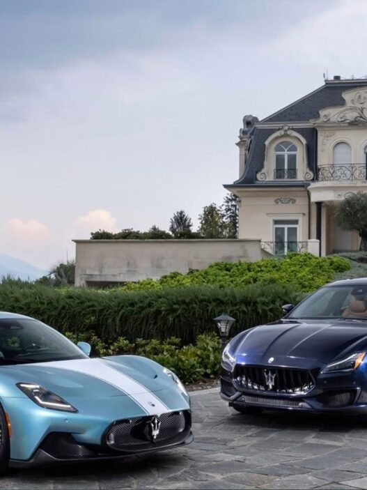 Dos coches de lujo de la maca Maserati, ambos de color azul, aparcados en un jardín frente a una casa muy elegante en un día nublado.