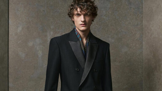 Modelo masculino con traje negro de doble botonadura, apoyado en un taburete alto en un entorno minimalista de tonos grises.