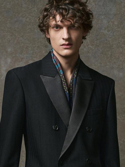 Modelo masculino con traje negro de doble botonadura, apoyado en un taburete alto en un entorno minimalista de tonos grises.