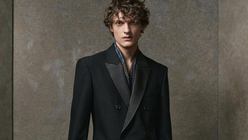 Modelo masculino con traje negro de doble botonadura, apoyado en un taburete alto en un entorno minimalista de tonos grises.