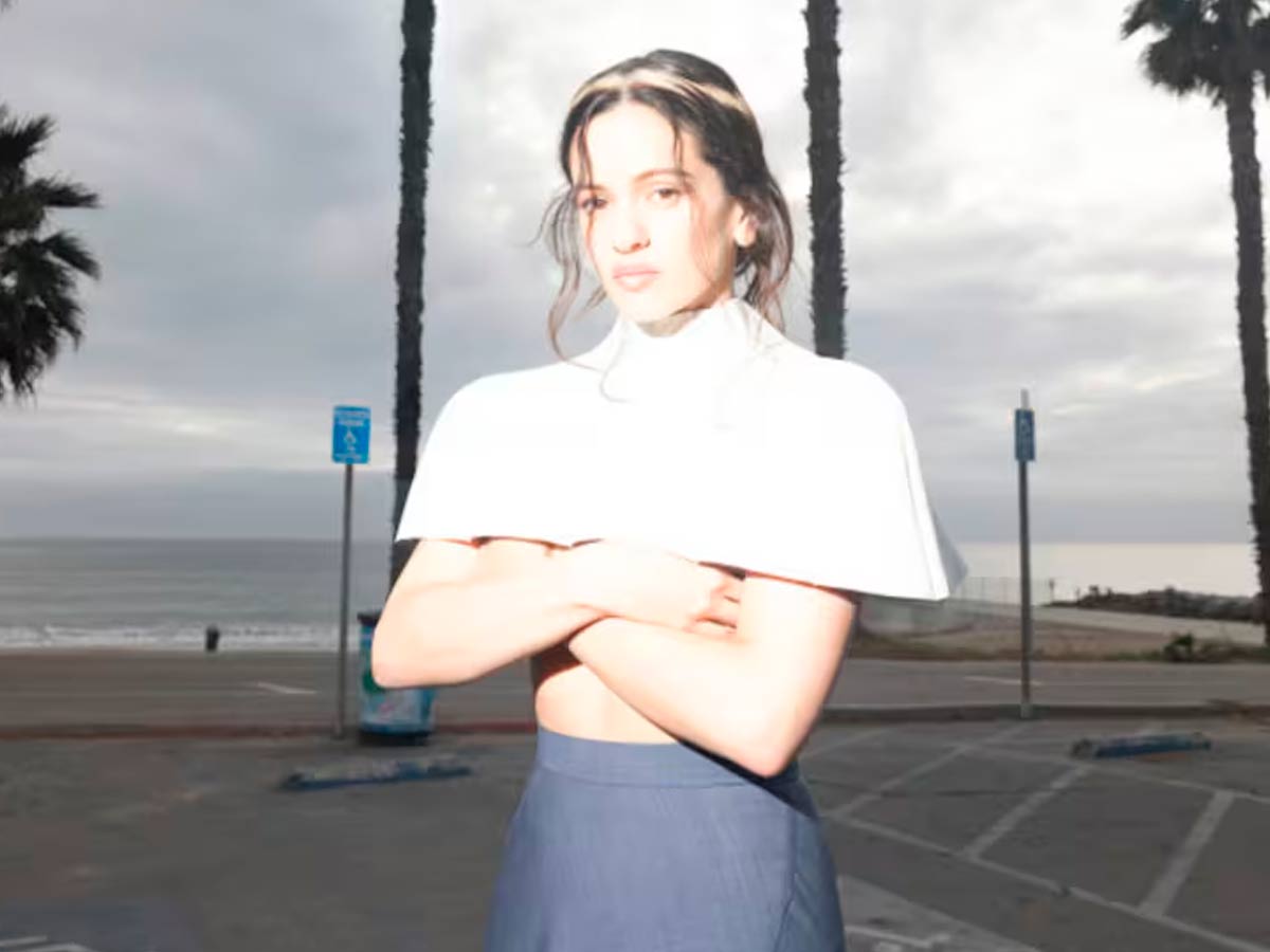 Imagen de Rosalía en una playa nublada, con dos palmeras de fondo y tapándose el pecho con las manos.
