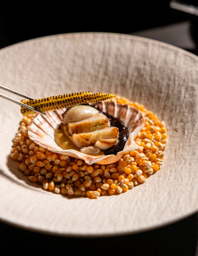 Vieira con crema de maíz y ajo negro del restaurante Tetsu, servida sobre granos de maíz tostado.