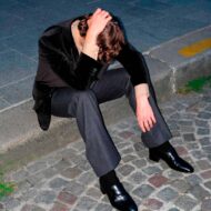 Las 5 marcas de ropa para hombre que más lo están petando y que en España aún no conoce nadie chico joven vestido muy elegante con un traje actual y moderno, a la última moda, sentado en la acera de la calle y tocándose la cabeza