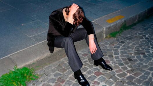 chico joven vestido muy elegante con un traje actual y moderno, a la última moda, sentado en la acera de la calle y tocándose la cabeza