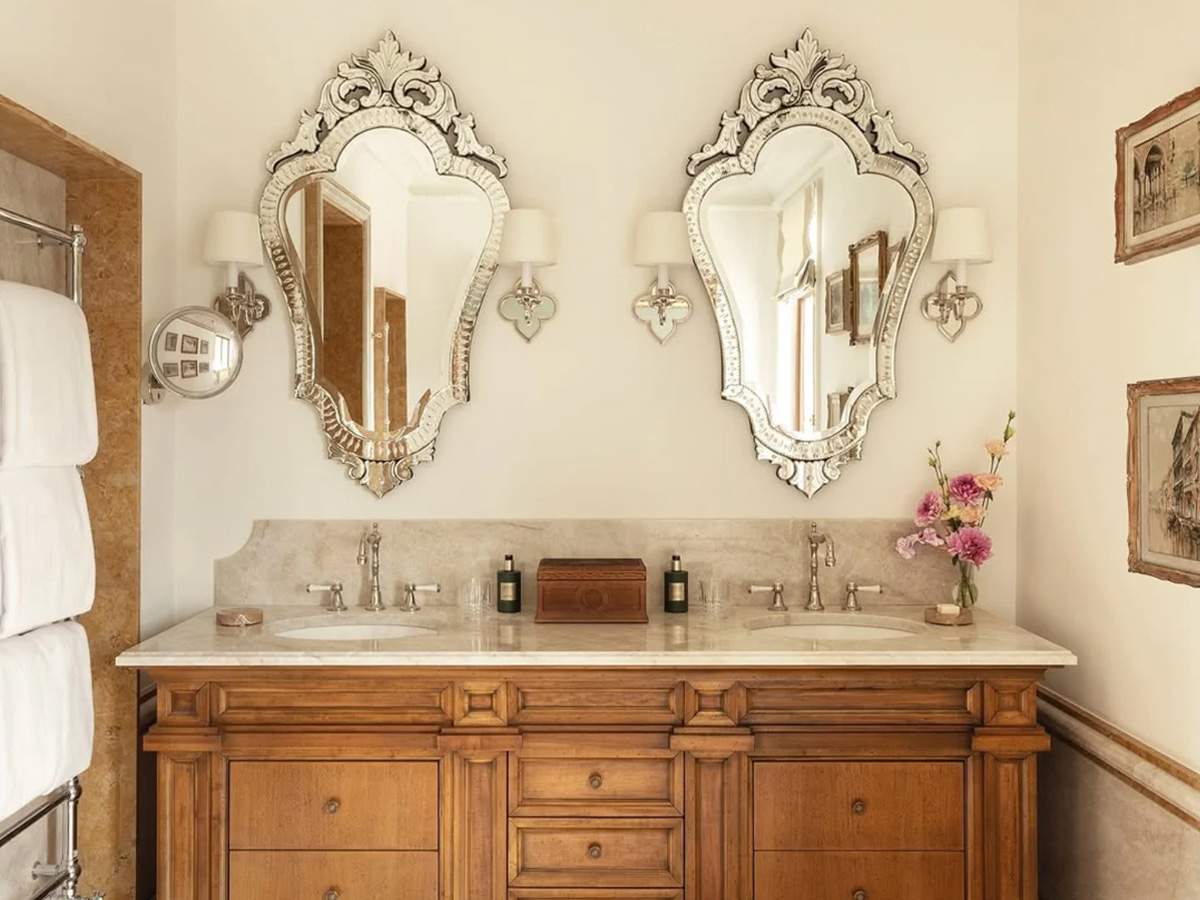 Baño de una de las suites del hotel Airelles Palladio Venezia con dos elegantes espejos y un lavabo estilo antiguo de madera y cuadros en las paredes