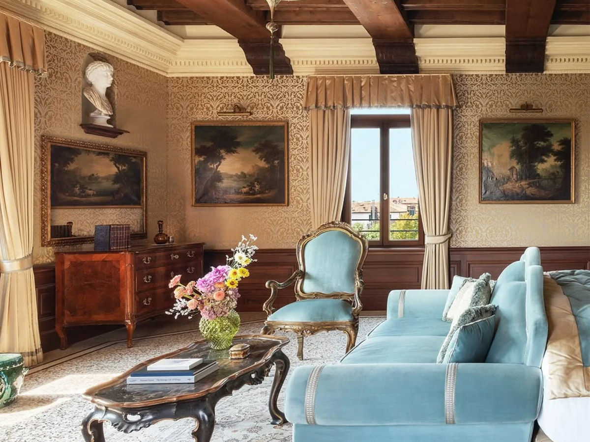 Suite del hotel Airelles Palladio Venezia, en la que se ve una habitación de techos de madera alto, con un elegante sofá tapizado en azul, frente a un enorme balcón con cortinas.