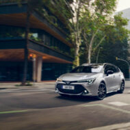 Qué coche comprar en 2026 si haces 30.000 km al año: 10 opciones que aguantan kilómetros de verdad Coche compacto Toyota Corolla híbrido de color gris circulando por una calle amplia de ciudad, visto desde el frontal en movimiento, con edificios modernos y árboles a ambos lados de la vía.