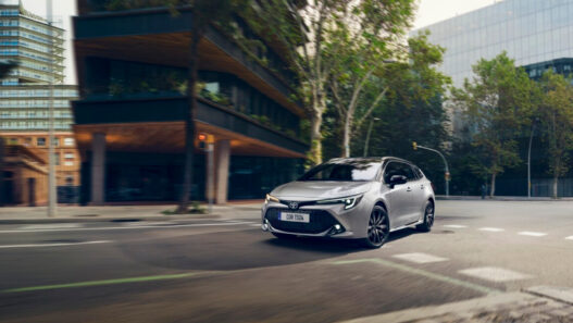 Coche compacto Toyota Corolla híbrido de color gris circulando por una calle amplia de ciudad, visto desde el frontal en movimiento, con edificios modernos y árboles a ambos lados de la vía.