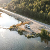 Las mejores rutas en autocaravana para descubrir España este 2026 Vista aérea de una furgoneta camper blanca estacionada junto a un lago o río, al lado de una carretera secundaria rodeada de bosque, con luz suave de última hora del día reflejada en el agua.
