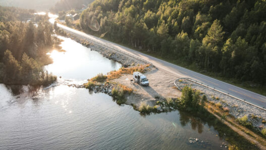 Vista aérea de una furgoneta camper blanca estacionada junto a un lago o río, al lado de una carretera secundaria rodeada de bosque, con luz suave de última hora del día reflejada en el agua.