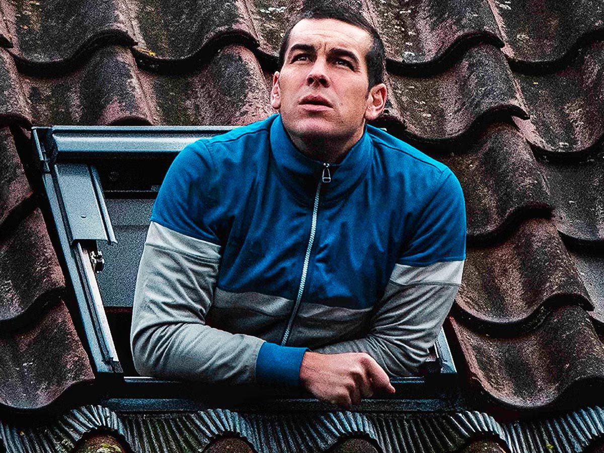 El actor Mario Casas asomado a la ventana de una boardilla, vestido con un chándal azul y gris, en una escena de la película "Muy lejos"
