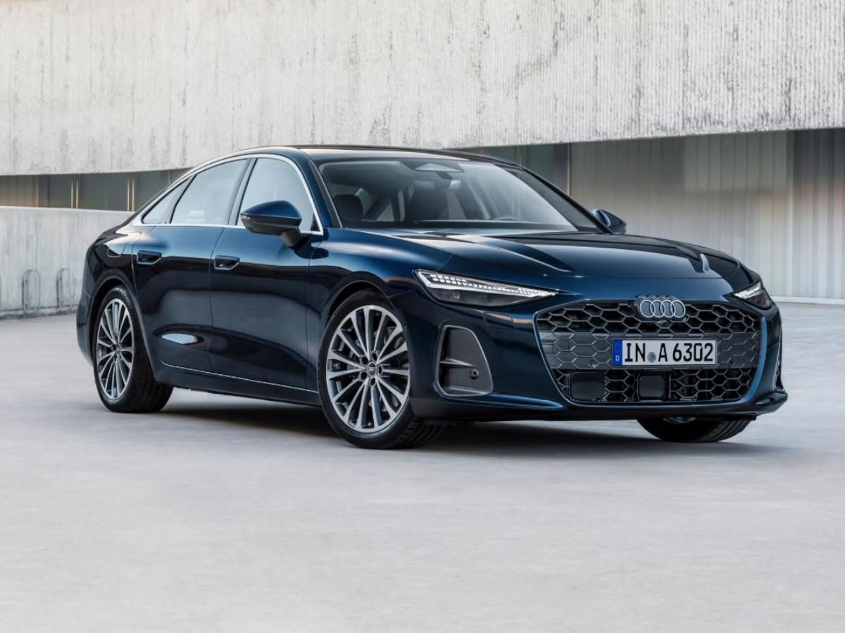 Berlina Audi A6 de color azul oscuro, aparcada frente a un edificio moderno de hormigón, vista desde el frontal y el lateral, con carrocería baja y líneas elegantes.